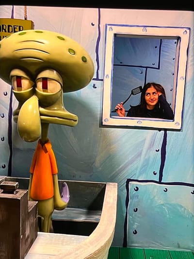 This is a photograph taken in Nijmegen, Netherlands, featuring a scene inspired by the animated television series SpongeBob SquarePants. In the foreground, a large, realistic prop of Squidward Tentacles is depicted, facing to the right and slightly upwards. His characteristic green body, large nose, and distinctive eyes are visible. He appears to be interacting with another character, likely Plankton, whose orange body and purple head are partially visible to the right of Squidward.
Above and behind Squidward, a square window is set into a wall painted to resemble the underwater environment of Bikini Bottom, with light blue and dark blue rectangular panels. Peeking out of this window is a young woman with long, dark hair. She is smiling and holding a black spatula, suggesting she is part of a theatrical or promotional event. Her expression is cheerful and engaging. The background wall is textured and painted in shades of light blue, with darker blue lines representing seams and circular black accents that mimic rivets or fasteners. To the far right, a portion of a green wall can be seen.
A yellow rectangular object with black text is partially visible on the left side of the frame, with the word "ORDER" clearly legible. The overall scene appears to be a set design, possibly for a stage production or an interactive exhibit, given the large-scale props and the presence of a person directly interacting with the set. The lighting suggests it is daytime, and there are no indications of specific weather conditions.