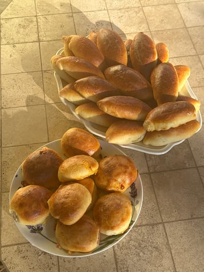 Two plates of baked goods are arranged on a tiled surface. The plate in the foreground is round and white with a floral pattern and contains several individual, golden-brown baked items that appear to be filled pastries. The plate in the background is a larger, white oval tray filled with numerous similar golden-brown baked items, many of which are elongated. The lighting suggests it is either early morning or late afternoon, casting long shadows across the tan and beige tiles. The background is out of focus, and no specific landmarks in Sharapovo, Russia, are identifiable.