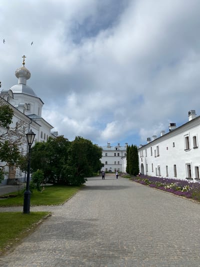 The image shows a view of the Valaam Monastery complex in Sortavala, Russia. In the foreground, a cobblestone pathway leads away from the viewer, bordered by green grass and mature trees on the left, and a white building with numerous windows on the right. Along the right side of the pathway, a flowerbed with purple and white flowers runs adjacent to the white building.

On the left side of the pathway, a white church with a golden dome topped with a cross is visible, along with a black lamppost. Further down the pathway, several people can be seen walking, and more white buildings characteristic of the monastery complex are visible in the distance. The sky above is cloudy with patches of blue sky. The overall scene depicts a serene and well-maintained courtyard within a historical religious site.