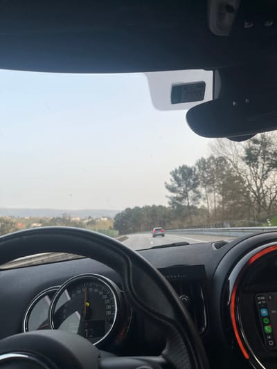 The image presents a first-person perspective from inside a moving vehicle on a multi-lane highway near Penafiel, Portugal. The vehicle's black, textured steering wheel occupies the lower left. Behind it, the instrument cluster displays a speedometer indicating approximately 120 km/h, alongside a digital readout showing "12" and "17:49." To the right, a circular air vent features an orange-red illuminated ring. Below this, an infotainment screen shows the time "17:49," a "5G" network indicator, and application icons. A black "VIRTUALS..." device is mounted centrally on the dashboard. The windshield exhibits minor dust and streaks. An electronic toll transponder or similar sticker is visible on the upper central windshield.
Ahead on the highway, a silver sedan with illuminated taillights travels in the same lane, which curves gently to the right. A metal guardrail borders the right side of the road, followed by a dense line of evergreen trees. To the left, a green embankment transitions to a panorama of distant cultivated land, scattered buildings, and rolling hills. The sky is uniformly hazy and bright, indicating an overcast or atmospheric condition. No individuals are explicitly visible within the vehicle.