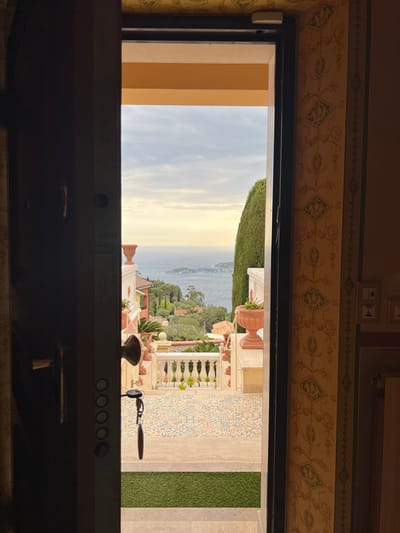 The image presents a view from inside a building, looking out through a doorway or large window onto a picturesque Mediterranean landscape in Èze, France. The interior features ornate beige and green patterned wallpaper with a decorative border. A dark frame surrounds the opening. On the left side of the interior frame, a white rectangular sensor or device is mounted. On the right, a light switch panel and another possible control panel are visible on the patterned wall.

Beyond the frame, the exterior scene unfolds under a clear sky with soft, diffused light, indicating either morning or late afternoon. A wide expanse of the Mediterranean Sea dominates the horizon, with a distant, verdant coastline. Closer to the building, a terraced outdoor area is visible, featuring white balustrades, terracotta planters filled with greenery, and a vibrantly patterned tiled path or patio. An area of artificial green turf is also seen on the right. A key on a keyring hangs near the bottom right of the doorway frame.

The dark structure forming the bottom edge of the interior view appears to be a counter or built-in unit, displaying five circular controls and a knob, along with a handle or lever, possibly suggesting a kitchen or service area. The overall setting depicts a luxurious and tranquil Mediterranean environment. No visible text is present in the image.