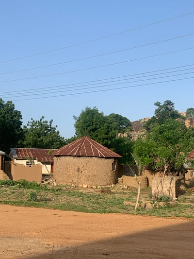 The image depicts a rural village scene in the city of Reak, Nigeria. The foreground is dominated by a dusty, orange-brown dirt road. Behind the road, a patch of dry, green ground is dotted with sparse vegetation. A cluster of traditional round houses with conical, corrugated metal roofs stands in the middle ground. The houses are constructed from mud and stone. To the left of the round houses, a rectangular building with a similar corrugated metal roof is visible. The background features lush green trees and rocky outcrops, with a clear blue sky above. Several power lines are strung across the sky. There are no people or animals visible in the image.