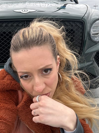 The image shows a woman with blonde hair, light skin, and rings on her fingers posing in front of a dark green Bentley. The grill and Bentley logo are visible. The location is unknown, and there is no indication that the location is in Dartford, United Kingdom. The woman is wearing a brown, furry jacket and grey hoodie. Her makeup includes eyeliner and shimmery eye shadow.