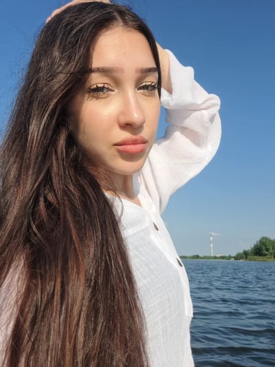 A young woman with long, dark brown hair is taking a selfie by a body of water. She is wearing a white, collared shirt with buttons. Her left hand is raised to her head, touching her hair. The background shows a clear blue sky, calm blue water, and a tree line with a distant smokestack. The scene appears to be outdoors on a sunny day.