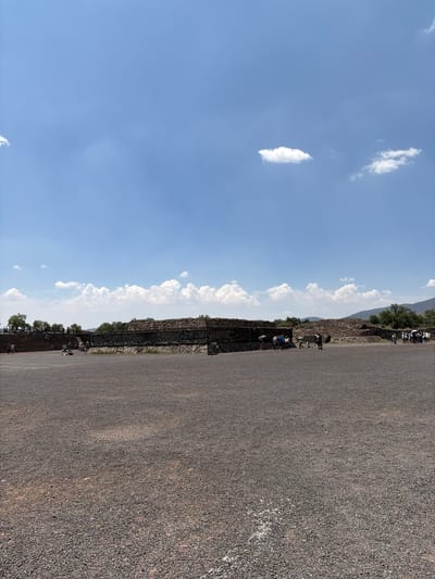 The image depicts a scene at Teotihuacan, an ancient Mesoamerican city located in the San Francisco Mazapa municipality, Mexico. The frame is oriented vertically with the sky occupying the left half and a ground surface, likely a pathway or part of a plaza, filling the right half.

The sky is blue with scattered white clouds. A linear stone structure, possibly part of a temple or wall, runs between the sky and ground. People are present, primarily concentrated along the stone structure. Vegetation, possibly trees, appears in the background. The ground is a rough surface, likely compacted earth or gravel.