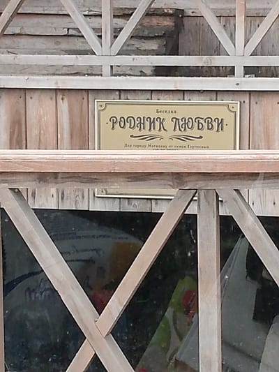 The image shows a sign on a wooden structure. The sign is golden with black lettering and has a decorative border. The text on the sign is in Russian. The top line reads "Беседка" (Gazebo). The main text reads "РОДНИК ЛЮБВИ" (Spring of Love). Below this, in smaller print, it says "Дар городу Магадану от семьи Гордеевых" (Gift to the city of Magadan from the Gordeev family). The background is a wooden wall, and in the lower part of the image, there is a dark reflective surface, possibly glass, showing blurred colorful shapes. The overall scene appears to be outdoors.