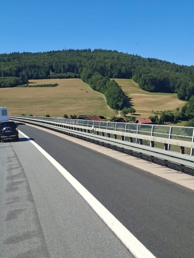 This is a photograph taken on a highway, likely in or near Riedenberg, Germany, on a clear, sunny day. The highway has multiple lanes, separated by a solid white line. On the left side of the image, a black SUV is parked or stopped near a white trailer. The SUV has a license plate that reads "KI-DJ 772".
The highway is bordered by a modern sound barrier, consisting of metal posts and clear panels. Beyond the sound barrier, the landscape features rolling hills covered with lush green forests. In the middle ground, there are open fields with dry grass and some scattered farm buildings with red roofs. A dirt road winds its way through the fields towards the buildings. The sky is a clear, bright blue. The overall impression is a rural highway scene on a pleasant summer day.