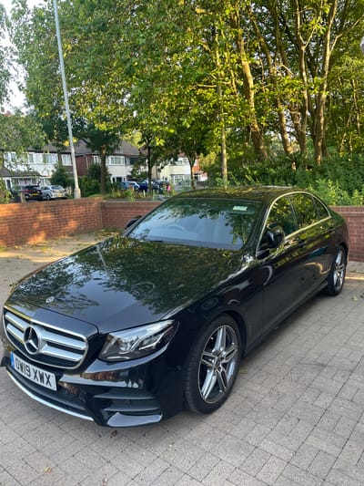 The image shows a black Mercedes-Benz E-Class parked on a paved sidewalk next to a brick wall. The car is clean and shiny, reflecting the surrounding environment. The license plate of the car reads "OW19 XWX". In the background, there are trees with green leaves, houses, and other parked cars. The sun is shining, casting shadows and creating a warm lighting effect, suggesting it is daytime, possibly late afternoon or early evening. The scene appears to be a residential street in Wolverhampton, United Kingdom, given the architecture of the houses and the style of the license plate. There are no people visible in the image. The overall atmosphere is calm and peaceful, depicting a typical urban or suburban setting.