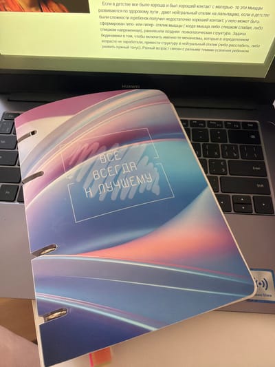 The image shows a close-up, overhead view of a laptop with a notebook partially covering the keyboard. The notebook has a colorful, abstract design with a quote in Russian that reads "ВСЁ ВСЕГДА К ЛУЧШЕМУ" (Everything is always for the best). The laptop screen is illuminated and displays text, but it is out of focus. The environment appears to be indoors, with a soft light source. There are no people visible in the frame. The image is taken from a slightly elevated angle, looking down at the objects. There is no visible information within the image to suggest a specific location within Кирпичного завода № 1, Russia.