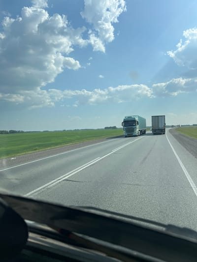 The image is a photograph taken from the driver's perspective of a vehicle traveling on a highway. The highway is paved with asphalt and has white lane markings. To the left of the highway is a vast, green field of crops. In the distance, a line of trees can be seen along the horizon. The sky above is blue with scattered white cumulus clouds.
Two large trucks are visible on the highway. The truck in the foreground is a light blue semi-trailer truck with a white cargo container. The truck behind it is a white semi-trailer truck with a gray cargo container. Several other vehicles, appearing to be cars and trucks, are visible further down the road in the distance, suggesting traffic.
The image appears to be taken in a rural or semi-rural area, given the expansive fields and open sky. The presence of a highway indicates a location with transportation infrastructure. The specific location of Chik, Russia, cannot be determined from the image alone, as the visual elements are generic to many highway landscapes in such regions. There are no people clearly visible in the image.