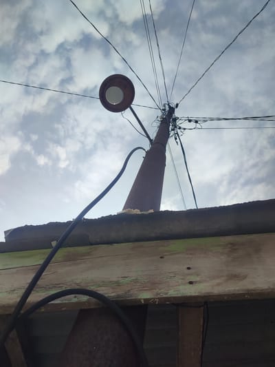 A low-angle shot shows a rusty, conical utility pole in Gampongjawa, Indonesia, supporting a round, overhead street lamp and a chaotic web of power lines against a cloudy sky. The pole emerges from behind the edge of a weathered wooden structure with peeling green paint and a rusty corrugated roof. A thick black cable arcs down from the street lamp, partially obscuring the lower portion of the image. The immediate foreground is dark, obscuring the objects it contains.