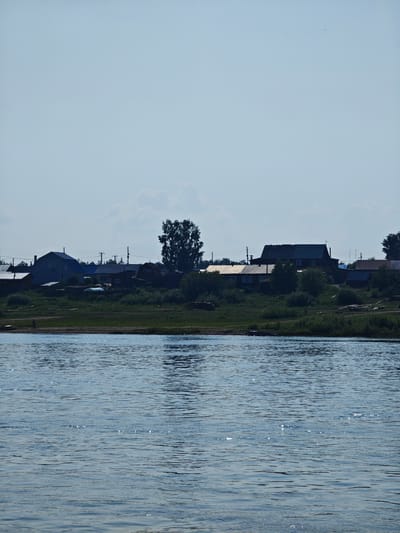 A wide, calm river with a slight ripple effect occupies the foreground of the image. The water is a muted blue-gray and reflects the light of the sky, creating shimmering highlights. In the middle ground, a densely wooded riverbank is visible, transitioning to a small village nestled on a slightly elevated area. The village consists of several houses with pitched roofs, some appearing to be wooden structures, while others are painted in lighter colors. Power lines and poles run along the edge of the village. A large, dark silhouette of a tree stands prominently to the right of the village. The background is a hazy, light blue sky with a few soft clouds. The overall impression is of a serene, rural landscape.