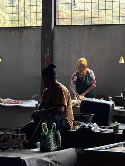 In an indoor market in Karangsambung, Indonesia, a woman wearing a yellow hijab and a green apron is shown from the chest up, preparing raw chickens at a counter. In the foreground, a person wearing a dark beanie and an orange t-shirt is seated facing away from the camera, looking towards the left. A woven green basket is placed next to them on the floor, and a vintage scale is visible on a surface to the left. The background features a large window with a grid pattern that allows natural light to enter, illuminating the dimly lit interior of the market. The overall scene depicts a moment of activity within a bustling marketplace.