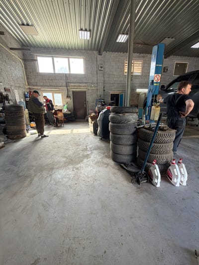 The image shows the interior of a car repair shop located in Yagul, Russia. Two men are present in the workshop. One man, wearing a green vest and brown pants, stands near a stack of tires, looking at his phone. The second man, in a red and black jacket, is behind him, appearing to be engaged in a conversation or activity. A car is positioned on a hydraulic lift in the background. The workshop floor is concrete and appears worn, with sunlight casting shadows from the windows and the ceiling. Various automotive parts, including multiple stacks of tires, oil canisters, and a hydraulic jack, are visible throughout the space, along with tools and equipment mounted on the walls. The walls are constructed of grey cinder blocks, and the ceiling is corrugated metal. A door is visible on the right side of the image, and windows are present on the far wall and the right side. A phone number is visible on the wall above the door.