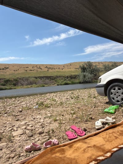 The image depicts a natural scene along a riverbank. The foreground is a dry, rocky terrain with sparse vegetation. Several pairs of footwear are scattered on the ground: pink flip-flops and two pairs of sandals, one white and one green. A brown blanket is partially visible in the bottom right corner. In the middle ground, a river flows, bordered by a steep, eroded embankment with greenery and shrubs. The background features rolling hills covered in dry grass and scattered bushes. A white vehicle is parked on the right side of the frame, its front fender and wheel visible. The sky above is blue with wispy white clouds. The scene suggests an outdoor recreational setting, potentially near the city of Boraldai, Kazakhstan, given the arid landscape and vegetation type.