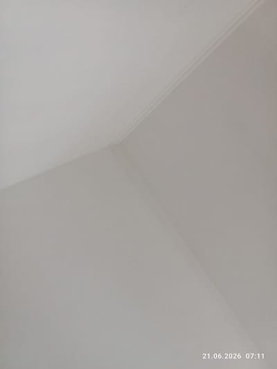 The image shows a light-colored textured wall meeting at a corner. A decorative molding runs along the edge where the two walls meet, with a thin, raised line above a recessed channel. The lighting appears to be diffuse, casting subtle shadows that highlight the texture of the wall and the details of the molding. The bottom right corner of the image contains a timestamp: "21.06.2026 07:11". No people or other objects are visible in the frame. The scene is likely an interior shot within a building in Жаңақұрылыс, Kazakhstan.