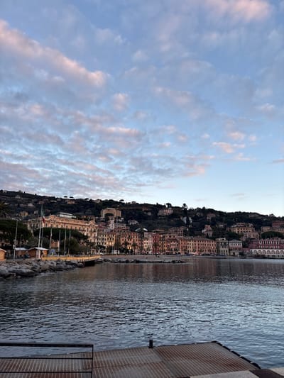 A picturesque view showcases a vibrant hillside town nestled along the coastline. Buildings cascade down the slope towards the water, reflecting a soft glow. To the left, the sky is filled with intricate patterns of clouds, tinged with faint colors of the setting sun. The water appears calm, with gentle ripples adding texture to its surface. On the right, part of a textured wall can be seen.