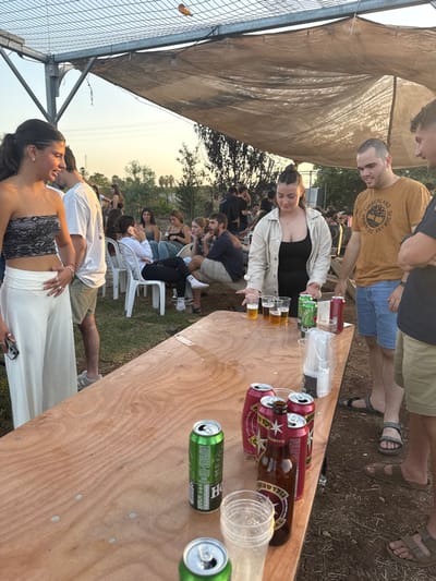 An outdoor social gathering is observed in the Gilboa Regional Council, Israel, under a light-colored tarp providing shade, supported by a metal frame. The scene is illuminated by late afternoon or early evening light. In the immediate foreground, a long, light-colored wooden table occupies the lower half of the frame. On the table, various beverage containers are present, including multiple green Heineken beer cans (some open, some stacked), red Goldstar beer cans, and an empty brown Goldstar beer bottle. Several clear plastic cups, some containing a light-amber liquid consistent with beer, are arranged on the table, along with a stack of unused clear plastic cups. Visible liquid spills and marks are on the table surface.
To the left of the table, a young woman with dark hair, wearing a black lace strapless top and white wide-leg pants, stands with her right hand resting on her abdomen, looking towards the left with a slight smile. To the right of the table, a young woman with dark hair tied back, wearing a beige jacket over a black top, stands facing the table with her hands positioned over the plastic cups containing beer. To her right, a young man wearing an orange "Timberland" t-shirt and blue shorts stands, looking to the left. Partially visible on the far right is another young man in a dark t-shirt and khaki shorts, also looking left.
In the mid-ground and background, numerous other individuals, primarily young adults, are gathered. Some are seated on white plastic chairs around smaller tables or on the ground, while others stand and socialize. Visible background elements include green foliage, trees, and power lines against a clear sky. The ground appears to be a mix of dirt and sparse grass.
