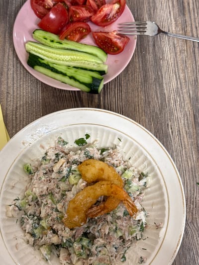 The image shows a close-up, overhead view of two plates of food on a wooden surface. To the left, a pink plate holds sliced tomatoes and cucumber sticks. A fork rests on the edge of this plate, with its tines pointing towards the wooden background. To the right, a larger white plate contains a salad, likely tuna or chicken salad, mixed with chopped vegetables and herbs. Two pieces of fried shrimp are placed on top of the salad. The overall scene appears to be a mealtime setting. The lighting suggests it could be daytime, possibly indoors given the lack of strong shadows. There is no visible text in the image. The location is Sunzha, Russia.