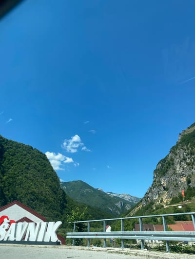 The image shows a scenic view of a mountainous region in Šavnik, Montenegro. In the foreground, a large white sign with red lettering spells out "SAVNIK." The sign is situated on the side of a road, with a metal guardrail in front of it. Behind the sign, a steep, tree-covered mountainside rises dramatically.
To the right of the sign, the road curves into a valley. The valley is flanked by steep, rocky cliffs on one side and more densely forested mountains on the other. Several small houses with red roofs are visible nestled on the slopes of the valley. In the distance, a series of layered mountain ranges stretch towards the horizon, their peaks softened by the haze of a clear day.
The sky is a vibrant, cloudless blue, with a few wispy white clouds scattered across the upper portion of the frame. The overall impression is one of natural beauty and a remote, mountainous landscape. The lighting suggests it is daytime, likely mid-morning or afternoon, with the sun casting distinct shadows. No people are visible in the image.
