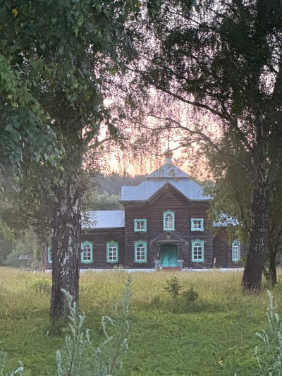 In Kushtomak, Russia, a traditional wooden church stands prominently amidst a natural landscape. The church, constructed from dark timber, features a metallic grey roof topped with a small golden dome. Its windows are adorned with bright green frames, and a matching green double door is visible at the entrance. The building is surrounded by tall, lush grass and is partially obscured by the branches and leaves of surrounding trees. The sky behind the church is illuminated by a soft, hazy sunset, casting a warm glow over the scene. The overall impression is one of serene, rustic beauty.
