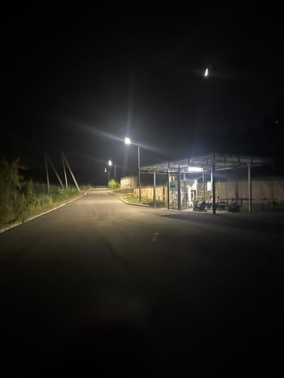 The image depicts an outdoor scene at night, likely in Uchterek, Uzbekistan. A paved road curves to the left, illuminated by streetlights that create lens flares. On the right side of the road, there is a covered industrial structure with pipes and machinery, possibly a gas station or processing facility. The area is surrounded by vegetation and a fence in the background. A sliver of the moon is visible in the dark sky. The overall impression is one of an industrial area at night, with limited visibility and artificial lighting.