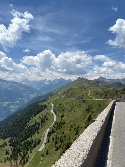 A scenic mountain road winds its way through lush green slopes dotted with evergreen trees. In the background, a range of majestic, snow-capped mountains pierces a bright blue sky filled with fluffy white clouds. The road is paved, with a concrete retaining wall visible in the foreground. No people or vehicles are readily apparent in the image, suggesting a serene and tranquil natural setting. The image captures a high-altitude landscape, likely within the Alpine region.
