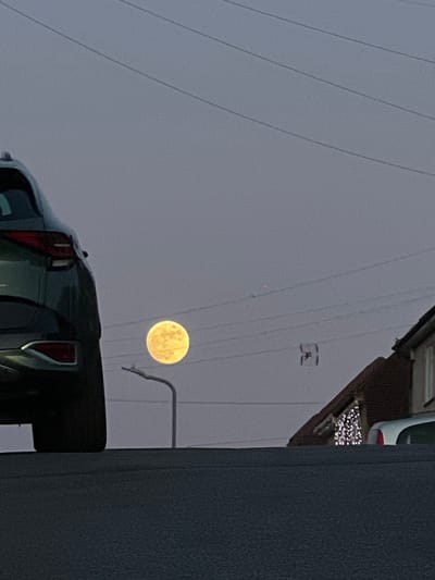 The image shows a full moon in a cloudy sky, partially obscured by power lines. A vehicle, possibly a pickup truck, is visible in the foreground, with its rear lights reflecting. Part of a building is seen on the right and top right. There are Christmas lights on top of the building. A street lamp is seen in front of the moon. The location of the image is High Wycombe, United Kingdom.