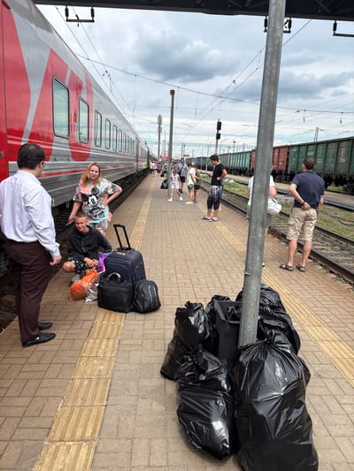 The image depicts a railway platform in Povorino, Russia, on a cloudy day. A red and silver passenger train is parked on the tracks to the left, with its doors closed. Several people are visible on the platform. In the foreground, a man in a white shirt and brown pants is talking to a young man who is crouching down next to several pieces of luggage, including a large blue suitcase, a black briefcase, and a black backpack. A woman in a Mickey Mouse t-shirt stands behind the crouching man. To the right of the platform, several black trash bags are piled up around a metal pole. Further down the platform, other passengers are seen standing and walking, some with luggage. In the background, freight trains are parked on adjacent tracks. The platform itself is paved with yellow and gray tiles, with a tactile warning strip running along the edge. Overhead power lines are visible, characteristic of a railway environment.