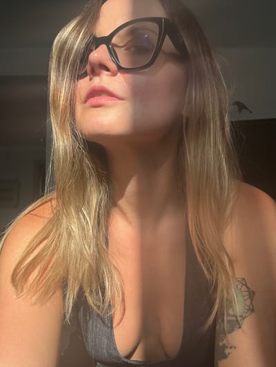 A close-up, waist-up shot of a woman wearing black-rimmed glasses and a dark grey top. She has blonde hair that is partially covering her face. The light from the right side of the frame casts a shadow across her face, highlighting her nose and upper lip. Her skin appears tanned, and there is a visible tattoo on her right shoulder blade. The background is dark and indistinct, suggesting an indoor setting. There is no visible indication of the location being Mesogi, Cyprus.