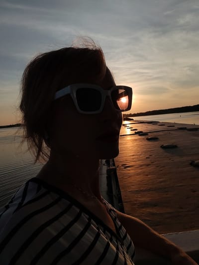 A silhouetted woman wearing large white-framed sunglasses poses for a selfie on a wooden pier at sunset. The sun is setting over a body of water, casting a warm orange glow on the scene. The woman's hair is light brown and windswept. She is wearing a striped top. The pier extends into the water and is lined with dark, rounded objects. The sky is a mix of blue and gray clouds. The location is identified as Shchetinino, Russia.