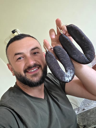 A male individual is depicted in an indoor setting in Suvorovo, Bulgaria, holding three dark-colored, U-shaped cured sausages in his raised right hand. The man has short, dark hair, a full beard, and is smiling directly at the viewer. He is wearing a dark olive green V-neck t-shirt. Each sausage is looped with a thin white string at its ends and exhibits patches of white mold on its surface. The background consists of a plain, light-colored wall, with a white electrical fixture visible on the upper left. A dark cord and a dark tiled surface are partially visible in the lower right quadrant of the image.