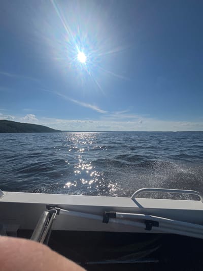 The image captures a bright, sunny day from the perspective of someone on a boat. The sun is a prominent, starburst-like feature in a clear blue sky, casting a lens flare that colors the sky with rainbow hues. Wispy clouds are scattered across the upper portion of the sky. The boat's railing, made of white material and a chrome handle, is visible on the right side of the frame, along with some dark interior of the boat.
The water is choppy with sunlight glinting off the waves, creating a sparkling effect. The wake of the boat is visible, with water droplets splashing. In the distance, a tree-covered shoreline is faintly visible, suggesting a lakeside or coastal setting. The image is oriented vertically. The overall mood is serene and evokes a sense of outdoor activity on a pleasant day. The time of day appears to be midday, given the intense sunlight. No text is visible in the image.