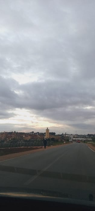 The image captures an outdoor scene at either sunrise or sunset, viewed from a slightly elevated vantage point. The sky is predominantly cloudy, featuring a mix of grey and white clouds, with a bright, warm yellowish glow emanating from the sun positioned low on the horizon.
Below the cloudy sky, a distant urban landscape unfolds, revealing a collection of buildings. A distinctive, lighter-colored building stands out amidst the others, illuminated by the sun's low light. Sparse greenery lines the horizon, separating the distant structures from the foreground.
In the midground and foreground, a wide, dark-grey paved road or track stretches into the distance, marked with faint lighter lines or boundaries. Along the left edge of the road, a dark, silhouetted figure, possibly a person on a bicycle or walking, is visible. Further along the road, in the distance, a few vehicles can be discerned, including what appears to be a small red car.
The overall lighting is soft and subdued, characteristic of dawn or dusk under overcast conditions. The scene appears to depict the outskirts of a town, aligning with the provided location context of Sidi Slimane Moul Al Kifane, Morocco. There is no visible text in the image.