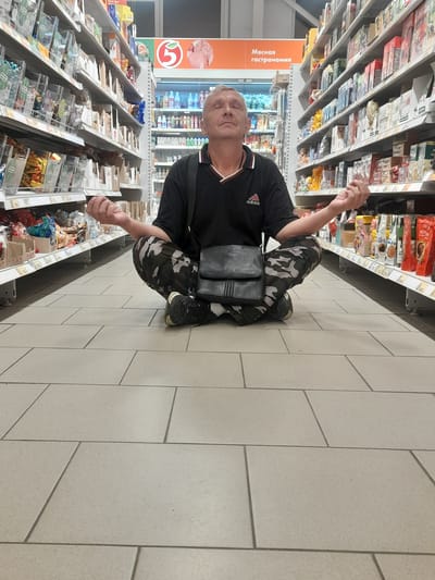 A middle-aged Caucasian male with grey hair is meditating in a lotus position on the floor of a brightly lit supermarket aisle. He is wearing a black polo shirt with the Adidas logo and camouflage pants. A black crossbody bag is slung over his shoulder. His eyes are closed, and his hands are resting on his knees, palms up. The shelves on either side of him are stocked with various food products. In the background, there is a refrigerated display case with drinks and a sign that reads "Мясная гастрономия" (Meat Delicatessen). The floor is tiled with large, light-colored square tiles. The scene appears to be in the city of Salsk, Russia, based on the context provided.