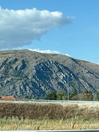 An elevated view shows a rugged, rocky mountain face dominating the upper portion of the frame. The mountain's slopes are streaked with lighter-colored rock formations and interspersed with patches of dry vegetation. A clear blue sky with scattered, wispy clouds stretches across the top. In the middle ground, a flat expanse of dry grass and scrub brush is bordered by a metal guardrail. Behind the guardrail, a few scattered evergreen trees are visible, along with a small building with a reddish-brown roof. A tall, thin pole, possibly a light pole or antenna, stands vertically in front of the mountain. The foreground consists of more dry grasses and some lighter green foliage. The image appears to be a landscape scene captured during daylight.