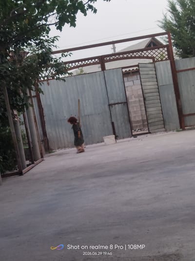 A young child with reddish-brown hair is standing in a concrete courtyard, facing away from the camera. The child is wearing a dark, possibly green, patterned dress and appears to be looking down at something on the ground. The background consists of a tall, corrugated metal fence with a wooden lattice structure above it. To the left, there is a leafy tree with thick branches. To the right of the child, a doorway is visible, framed by white brick and a section of metal siding. The ground is a rough, light-colored concrete surface. The overall environment suggests an outdoor urban or semi-urban setting. The time stamp on the image indicates it was taken in Bokhtar, Tajikistan on June 29, 2026, at 19:46.