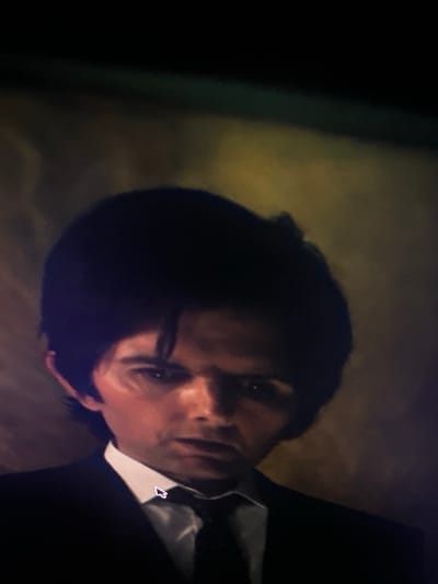 A still image, likely captured from a digital display, depicts a close-up portrait of an adult male, observed in Maisons-Alfort, France. The subject, positioned centrally, has dark, parted hair, a pale complexion, and dark eyes. He is attired in a white collared shirt, a dark tie, and a dark suit jacket. His facial expression is serious, with a direct and intense gaze. The background is dark, indistinct, and out of focus, exhibiting muted tones of yellow and green. A white computer mouse pointer is visible on the subject's tie, positioned to the left of the knot.