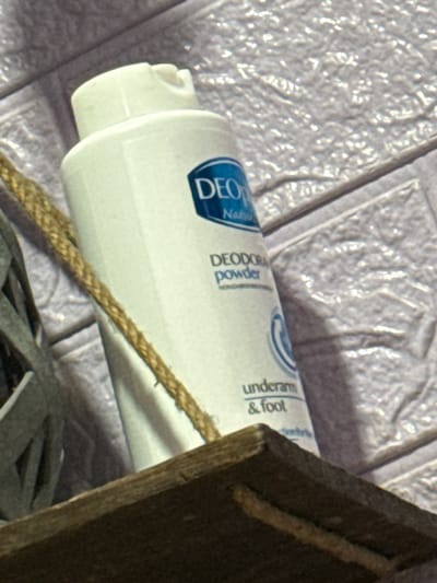 This is a close-up, low-angle shot of a white bottle of deodorant powder, possibly for underarms and feet, lying on a textured, light-colored surface. The bottle has blue and white labeling with text that is partially visible. The word "DEODOR" is clearly legible on the bottle, along with "powder" and what appears to be "underarm & foot". To the right of the bottle, a rough, textured, dark brown object, possibly a piece of wood or bark, is positioned diagonally, partially obscuring the bottle. A braided, tan-colored cord runs diagonally across the foreground, from the lower left to the upper right, further obscuring the view of the bottle and the textured surface. The lighting is harsh, creating strong highlights and shadows, suggesting an indoor setting, possibly with artificial light. There are no people visible in the image, and the scene appears to be a still life arrangement rather than capturing an active event. The overall impression is one of texture and detail focusing on the objects themselves. The location context of Manila, Philippines, does not offer any specific visual cues within the image itself.