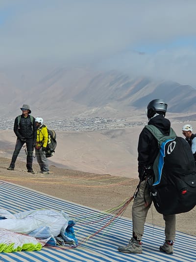A wide-angle, medium shot captures several people preparing to paraglide from a launching area above Alto Hospicio, Chile. The scene is dominated by the mountainous landscape and the city sprawling below. On the left of the frame, a bank of clouds partially obscures the horizon.

Three individuals are in focus, seemingly readying for flight. The person in the foreground wears a black quilted jacket, gray pants, and dark shoes, with a wide-brimmed hat and sunglasses. Behind them, a second person wears a yellow jacket and gray pants, with a white helmet. A third individual is oriented toward the top of the frame, clad in black and gray with a blue accent on their jacket, and a dark helmet.

The launching area is marked with blue and white stripes. The ground slopes away from the takeoff point, revealing a dry, brown landscape and the city of Alto Hospicio in the valley below. The paragliding equipment, including the colorful control lines and inflated parachute, are visible on the right side of the frame.
