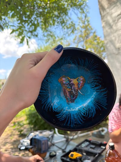 A close-up shot shows a hand holding a dark blue bowl with an image of a vibrantly colored elephant and the word "Thailand" in white script. The bowl has a radiating blue pattern. The background is blurred, showing green foliage, a blue sky, and a concrete structure, suggesting an outdoor setting. In the foreground, out of focus, is a toolbox with various tools and a brown bottle. A person in a pink and white striped shirt is partially visible to the right. The image is not specific to Komsomolskiy, Russia.