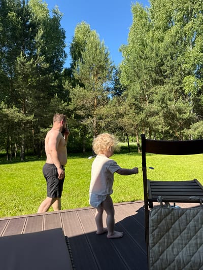 This image depicts an outdoor scene in Копнино, Russia, on a clear, sunny day. In the foreground, a dark slatted wooden deck extends across the bottom of the frame, with a partial view of a dark rectangular cushion or mat on the lower left. A small child, barefoot with curly blonde hair, stands on the deck in the right-center, facing away from the viewer. The child wears a light-colored t-shirt and a diaper, and holds a small, dark, spoon-like object attached to a string, extending it towards a dark wooden outdoor chair or table that partially obscures the child's lower body. A quilted, light-colored fabric item is hanging from the lower part of this wooden furniture.
Beyond the deck, a vibrant green lawn spans the midground. An adult male, shirtless with a beard and short dark hair, walks across the lawn from left to right, facing partially away from the viewer. He wears black athletic shorts and is holding a dark smartphone to his right ear, as if engaged in a call. A wedding ring is visible on his left hand.
The background consists of a dense treeline, featuring a mix of tall coniferous trees (pines or firs) and deciduous trees with lighter green foliage (birches). A clear, bright blue sky is visible above the treeline. The strong sunlight casts distinct shadows on the wooden deck.