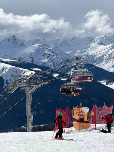The image depicts a winter ski resort scene in Lunnaya Polyana, Russia, under a partially cloudy sky. In the foreground, a snowy ski slope is visible. On the left, a skier wearing a reddish-brown jacket, black pants, a bright green helmet, and goggles stands on skis, holding ski poles. To the right, a snowboarder in a red jacket and black pants is bent over a snowboard lying on the snow, appearing to adjust equipment. Behind these individuals, a section of red safety netting encloses a large yellow snow cannon, identified with "T40" text, positioned on the snow.
In the midground, a ski lift system dominates the view. A tall, gray metal ski lift tower supports multiple cables traversing the scene. Two enclosed chairlift cabins are prominent on the cables; the upper cabin appears empty and displays an advertisement for "СБЕР Первый" with a QR code, while the lower cabin carries multiple passengers wearing winter attire. Dark green pine forests cover the lower slopes and valleys in the middle distance.
The background features a dramatic range of snow-capped mountains under a mix of clear sky and scattered white clouds. The peaks exhibit significant snow cover and rugged terrain.