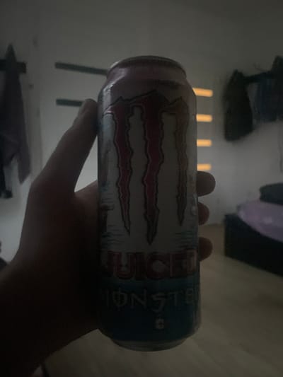 A close-up, dimly lit, indoor, eye-level, vertical shot shows a person's hand holding a can of Monster Energy drink. The can is mostly visible, with the red and white Monster logo prominently displayed. The text "JUICE" and "MONSTER" are also legible on the can. The background is blurred and indistinct, with hints of furniture and walls, suggesting a domestic setting. The overall lighting is low, creating shadows and obscuring some details of the environment.