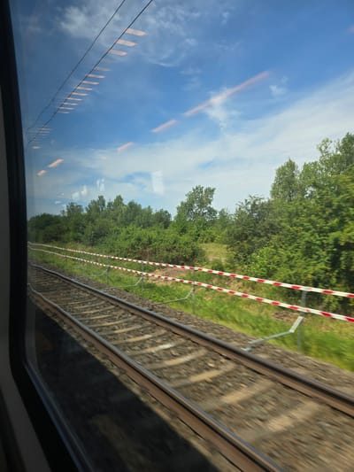 The image shows a view from a train window as it travels through a rural area, likely near Nordhausen, Germany. Twin train tracks run parallel, with ballast beneath them. A red and white striped barrier runs alongside the tracks. Lush green trees and bushes line the background. The sky is blue with scattered clouds, and reflections of interior lights from the train are visible on the window. The motion blur indicates the train is in transit.