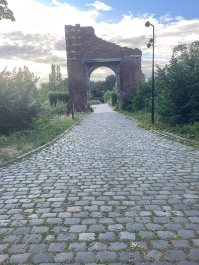 The image captures a stone-paved pathway curving through a green park-like setting in Weert, Netherlands. The pathway, laid with grey, rectangular stones interspersed with grass, dominates the foreground and recedes into the distance. To the left of the path, a crumbling brick archway stands as a historical remnant, possibly part of an old structure, with some supporting columns visible. The archway is weathered, showing signs of age with missing bricks and exposed stone. Beyond the arch, trees and foliage form a lush backdrop, with hints of more greenery and a clear sky with scattered clouds suggesting it's either morning or late afternoon. A street lamp with three lights is positioned near the archway, indicating a public space. The overall mood is serene and slightly melancholic, with the historical structure evoking a sense of the past within a natural, tranquil environment. No people are visible, and there is no visible text.