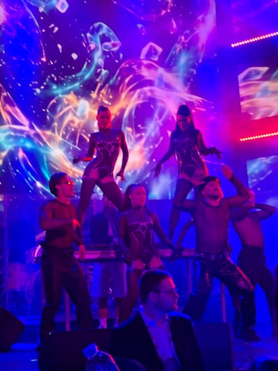 In Tsilkani, Georgia, a stage performance is underway under a vibrant, abstract digital backdrop that pulsates with blues, purples, and reds. Several performers are on stage, some wearing futuristic, form-fitting outfits with geometric cutouts and shimmering details. One female performer stands prominently on a raised platform, her pose dynamic and theatrical, while another is positioned lower, looking up and to the side. A male performer in glasses and a dark shirt stands in the foreground, seemingly observing the performance. Other individuals, likely musicians or crew, are visible behind the main performers, operating equipment such as keyboards and drums. The lighting is dramatic, casting strong colors and shadows across the scene, contributing to a high-energy, entertainment-focused atmosphere.