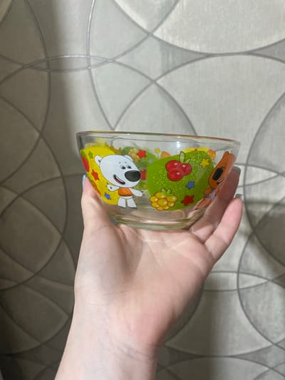 An adult's hand, likely female, holds a transparent glass bowl indoors. The hand has light-colored nail polish. The glass bowl is decorated with colorful cartoon characters, including a brown dog-like figure and a white bear-like figure, along with green patches, red berries, yellow berries, and multicolored stars. The white bear character has a black nose and ears, a red mouth, and wears an orange top, depicted smiling and with arms outstretched, seemingly floating on a yellow circle. The brown character is seen from above, wearing blue bottoms, and has an open mouth. The background consists of a wall covered in light-toned wallpaper, featuring a repeating geometric pattern of interlocking gray and white circles or ovals that create a textured, lattice-like design. There is no visible text on the bowl or in the background. The image is taken in Verkhnevesyoloye, Russia, showing an everyday domestic scene.