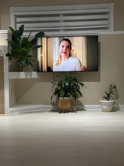 The image shows a flat-screen television mounted on a wall in what appears to be a living room in Springboro, United States of America. The television displays a woman with blonde hair wearing a white shirt. In front of the television, on a white shelf, is a large potted plant with broad green leaves. To the right of the television, on the floor, is another potted plant with long, slender green leaves in a wicker basket. Further to the right, on the floor, is a smaller potted plant in a white pot. The wall is painted a neutral color, and the floor is covered in light-colored wood-look planks. A white baseboard runs along the bottom of the wall. Above the television, there is a window with horizontal blinds. To the left of the television, a portion of a fireplace with a textured surface and decorative molding is visible. The overall scene suggests a domestic indoor setting.