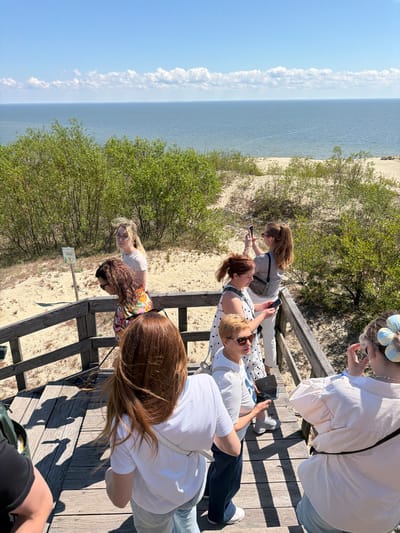 A group of people stand on a wooden boardwalk overlooking a sandy dune landscape and the sea under a blue sky with scattered clouds. A sign with an image of a person walking is visible on a pole to the left of the boardwalk. Several individuals are holding smartphones, suggesting they are taking pictures or videos. The people are dressed in casual summer clothing, and the overall atmosphere appears to be one of tourism or leisure. The location is Morskoye, Russia, likely a coastal area with sand dunes.